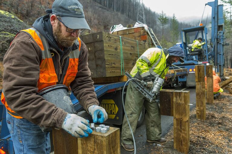 A road worker handles nuts and bolts on a timber post beside a truck during guardrail work